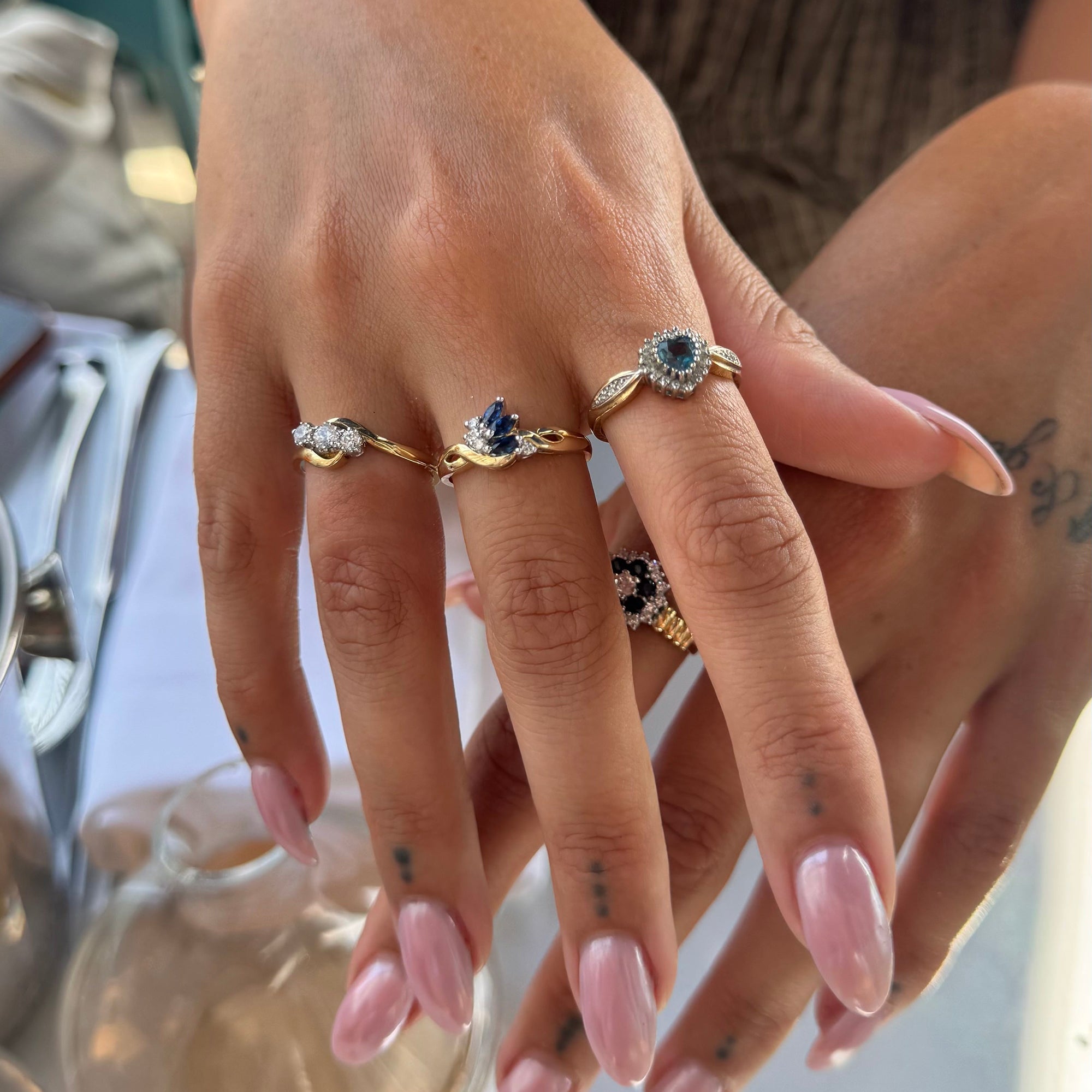 A close-up of a person's hands wearing four gold rings with blue and clear gemstones, set against a softly blurred background - seolgold