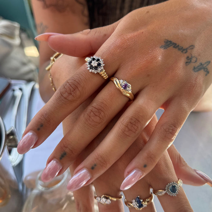 Close-up of a person's hands wearing multiple gold rings with different gemstone settings, including a black-and-white flower ring, a yellow stone ring, and smaller blue and white rings - seolgold