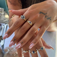 A close-up of a person's hands wearing multiple gold rings with floral and oval designs, some accented with white, blue, and purple stones. The nails are manicured in a light pink color, and the background is softly blurred - seolgold