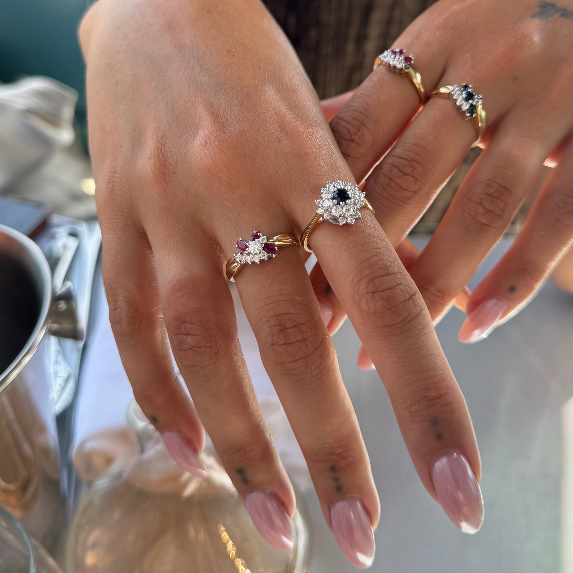 A close-up of a person's hands wearing four gold rings with floral diamond settings and colored gemstone accents, photographed in a real indoor setting with a mug and glass in the background - seolgold