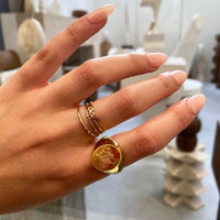 A close-up of a hand wearing a gold signet ring with an engraved letter B design and a small gold chain ring stacked on one finger, set against a softly blurred indoor background - seolgold