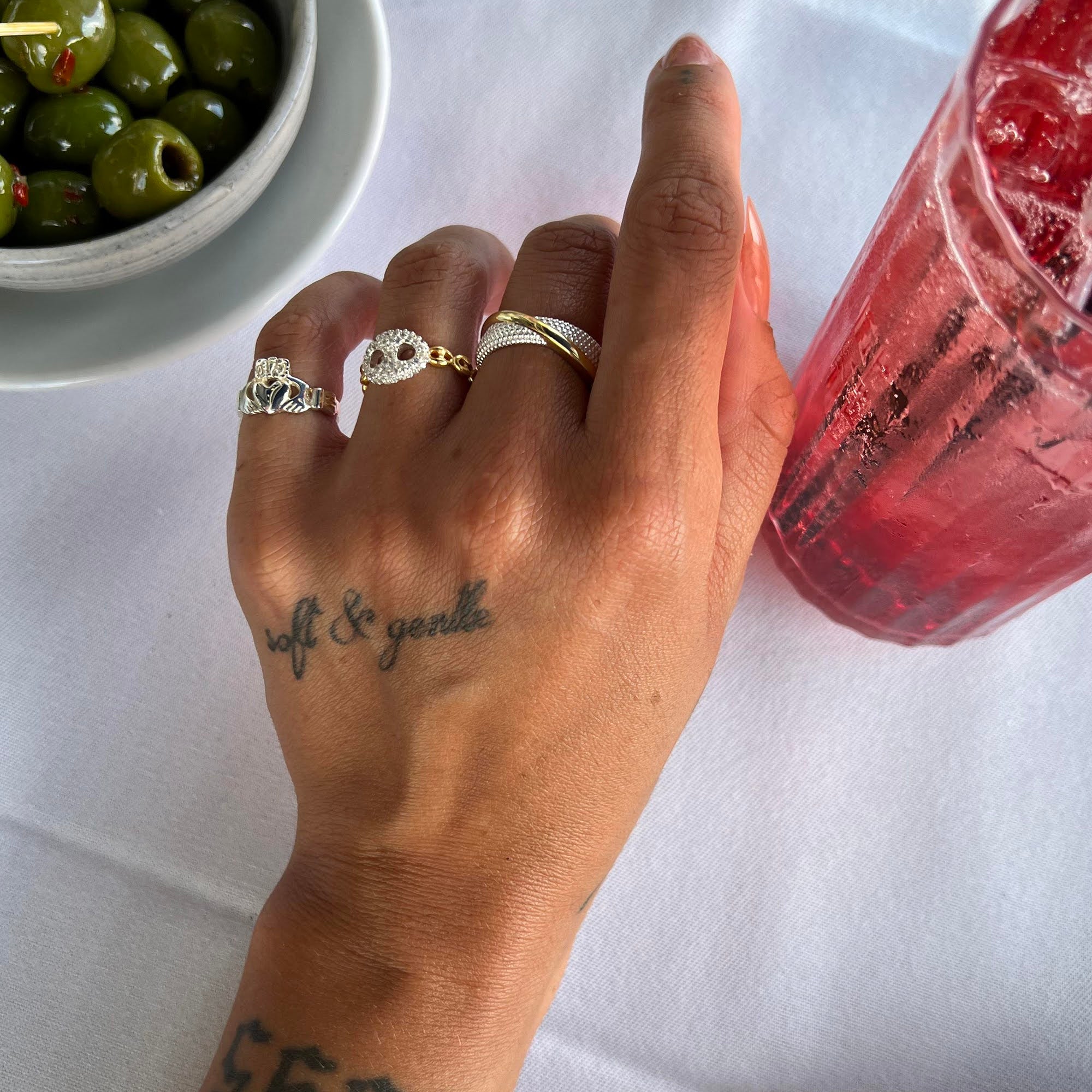 Hand with 'soft & gentle' tattoo holding a pink drink, with olives in the background - Seol Gold