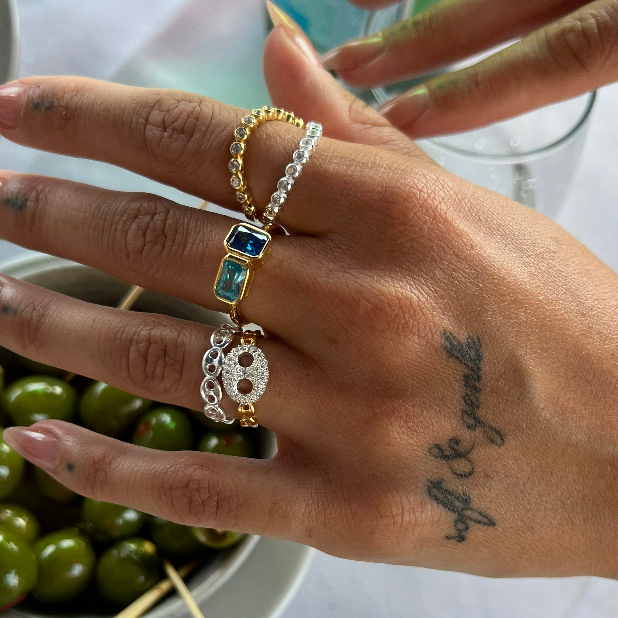 Hand wearing multiple rings with a tattoo on the wrist, holding green olives - seolgold