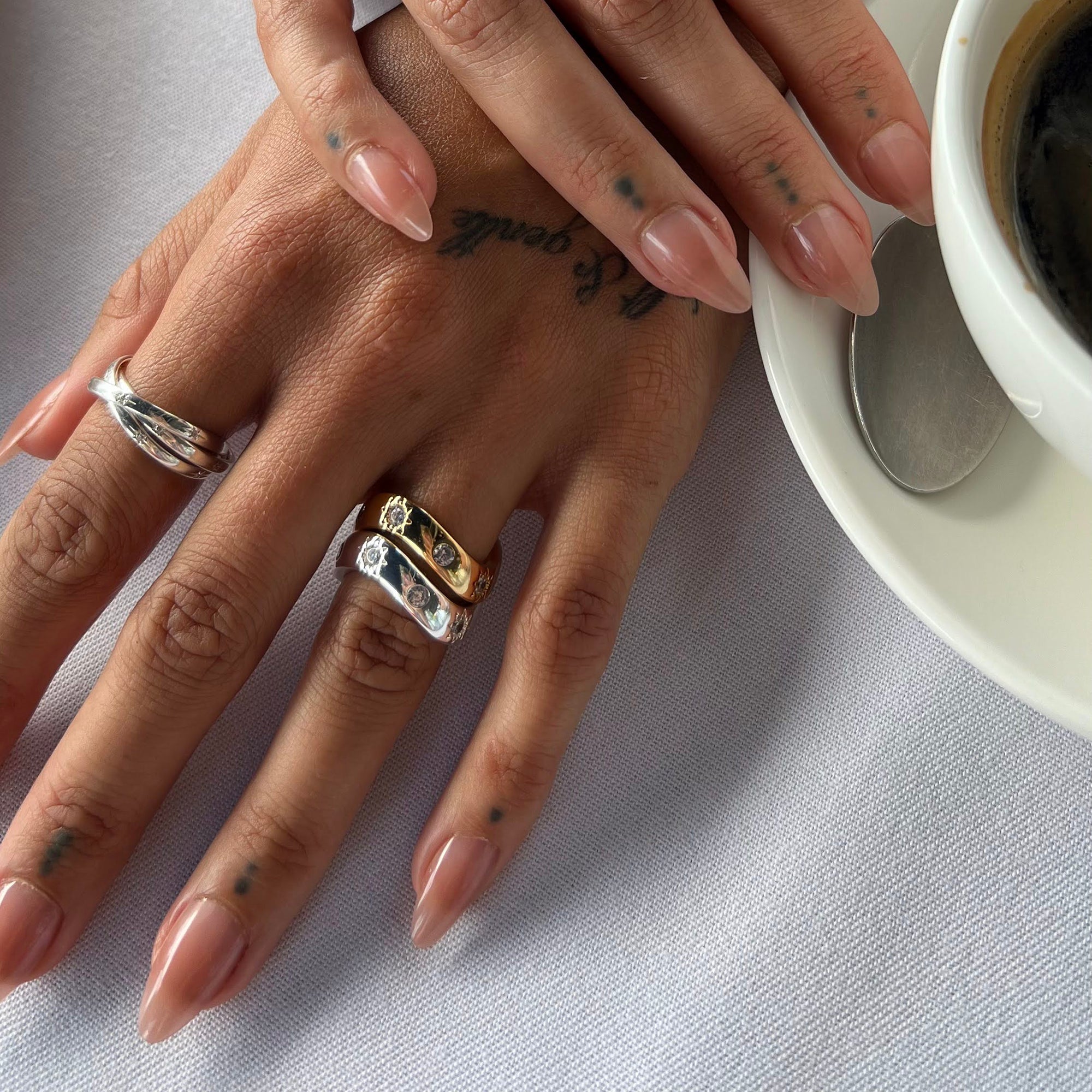 Close-up of hands with rings and tattoos, next to a cup of coffee - seolgold