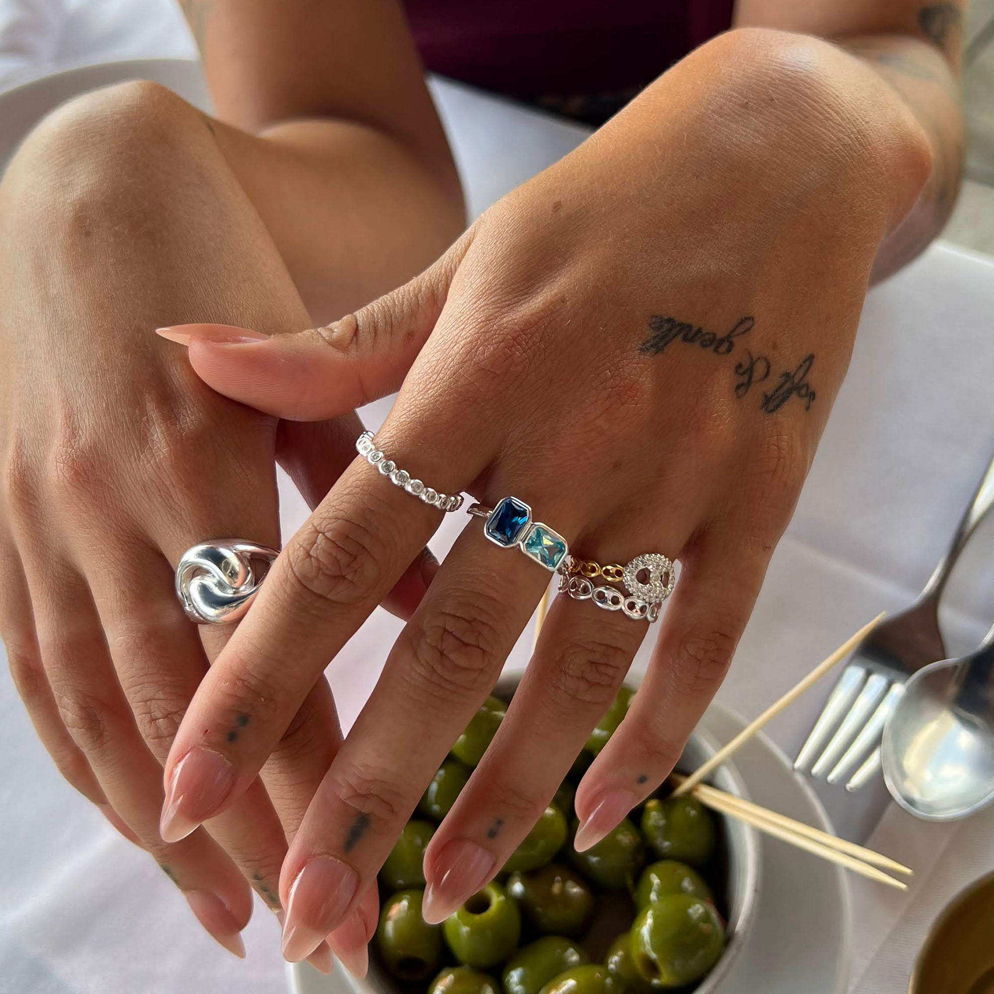 Close-up of hands with multiple rings and a tattoo, set against a blurred background with food and cutlery - seolgold
