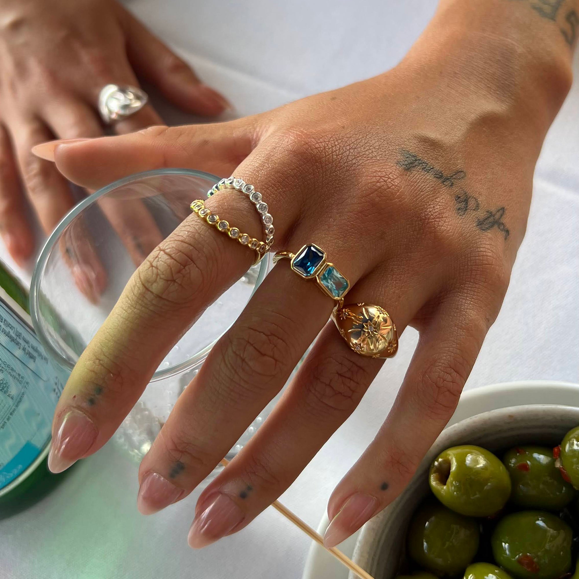 Close-up of a hand wearing multiple rings with a background of olives and a glass - seolgold