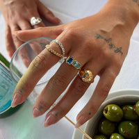 Close-up of a hand wearing multiple rings with a glass and bowl of olives in the background - seolgold