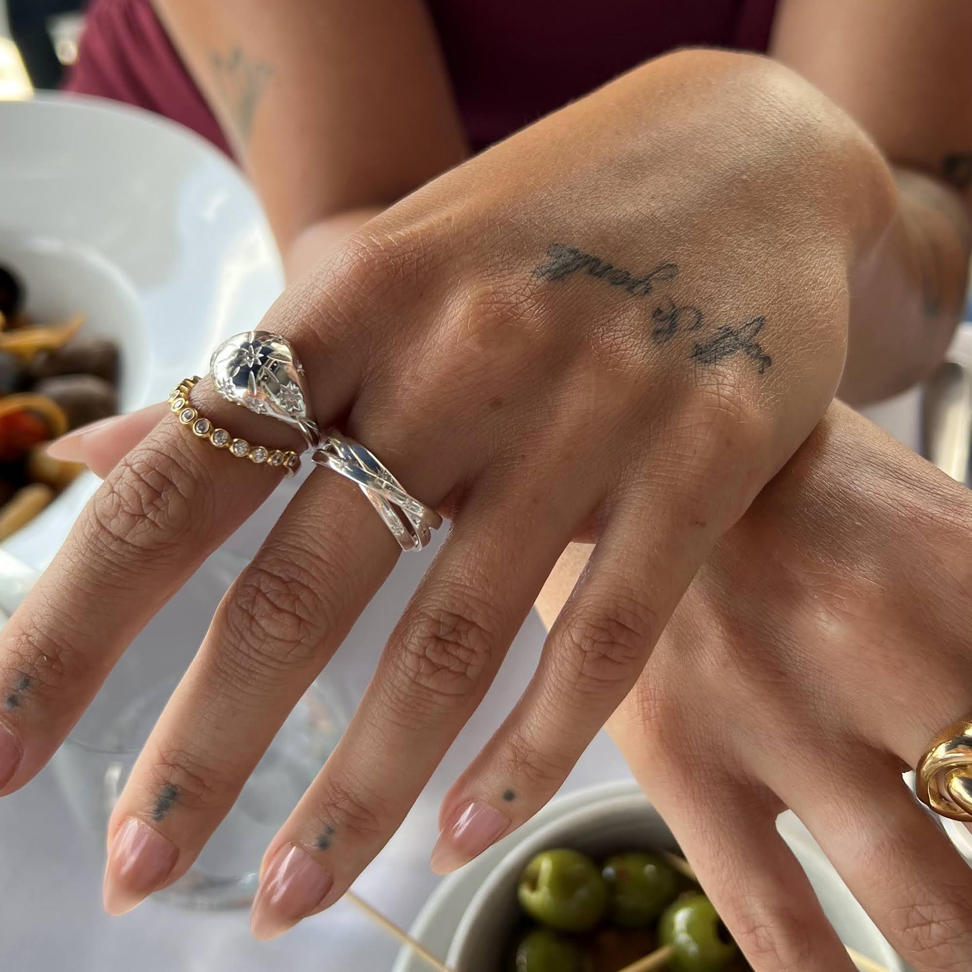 Close-up of a hand with multiple rings and tattoos, set against a blurred background - seolgold