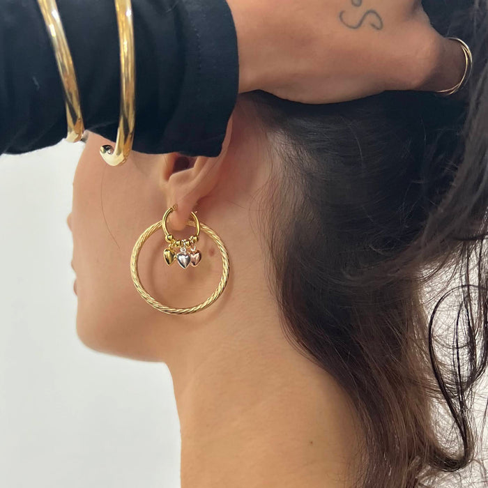 Close-up of a person wearing gold hoop earrings with heart charms on a white background - Seol Gold