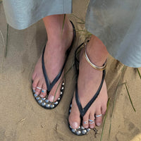 Black sandals with silver studs worn on a sandy surface wearing toe rings - seolgold