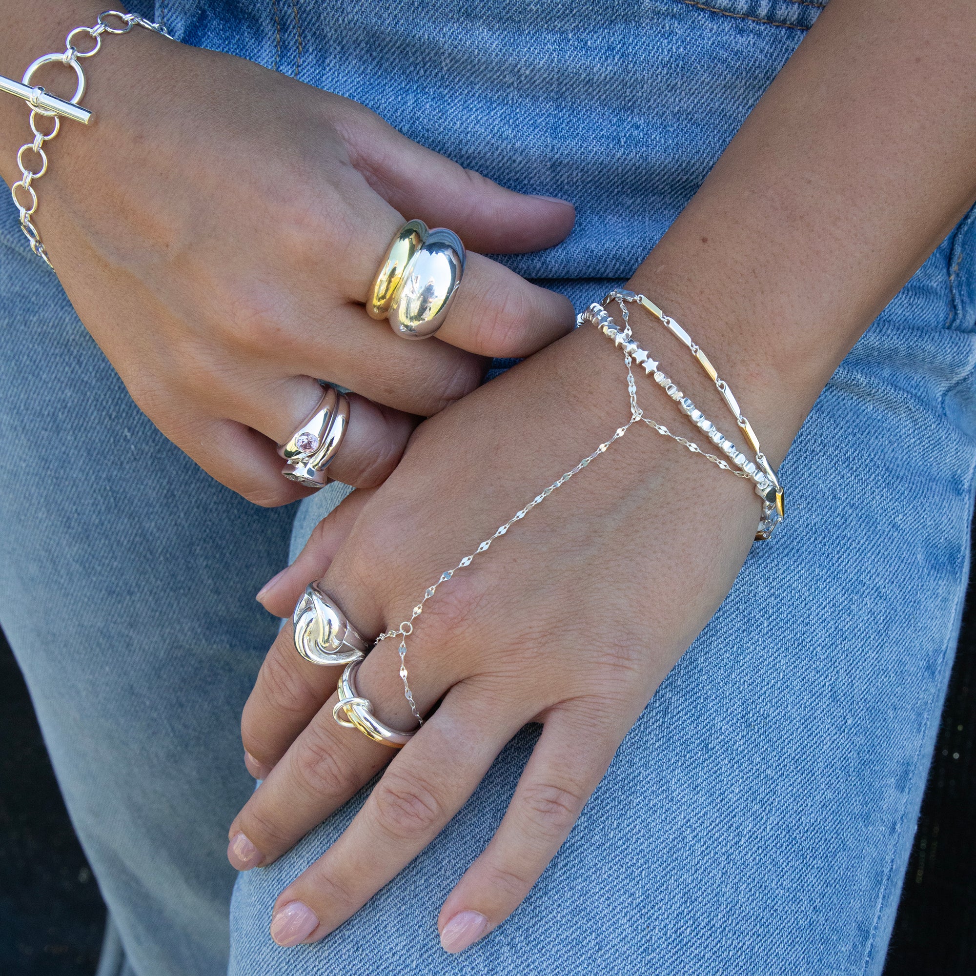 A close-up of a person's hands and lap wearing a delicate silver chain hand bracelet with a matching chain bracelet on the wrist, styled with multiple statement rings. - seolgold