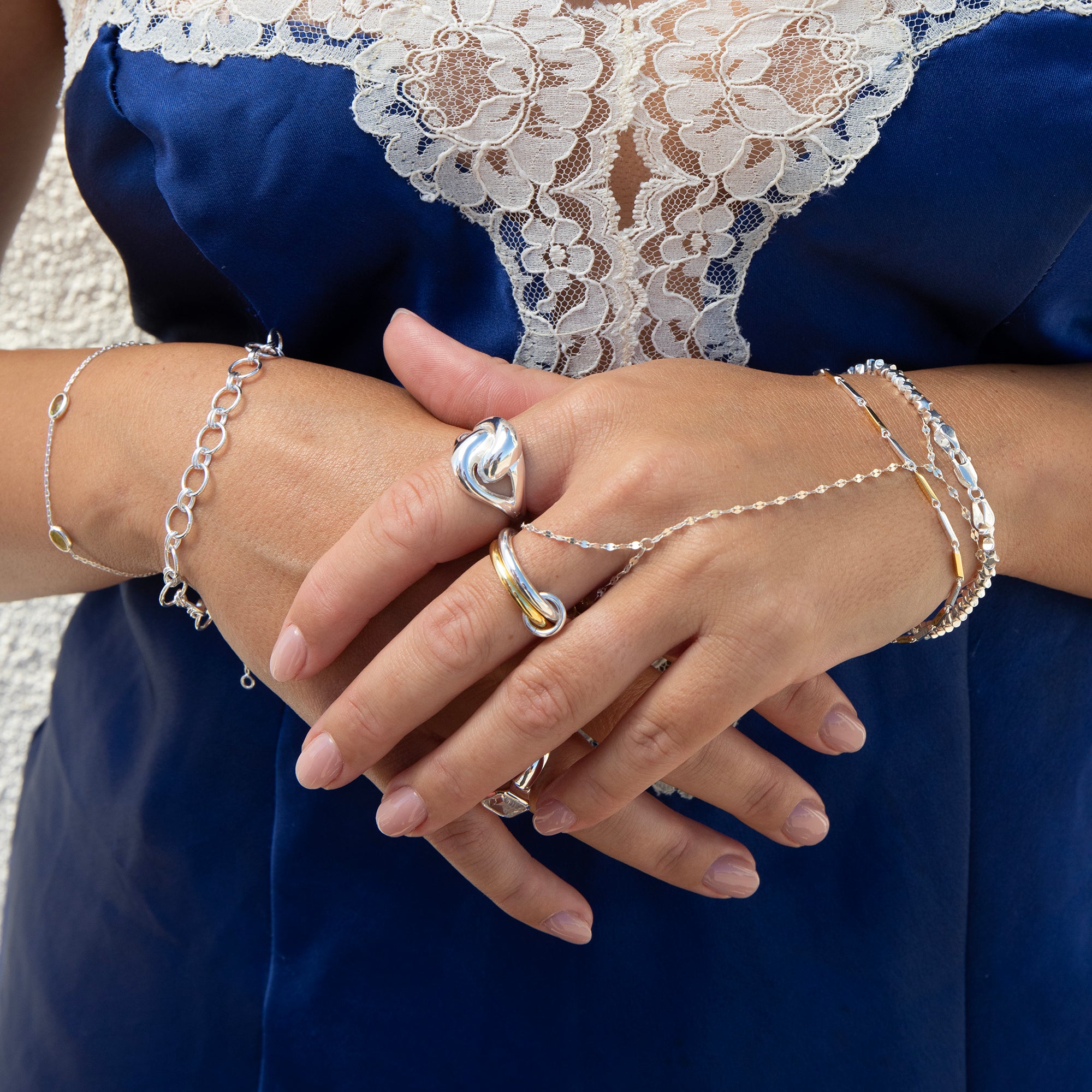 A close-up of a person's hands wearing multiple delicate silver-toned rings and chain bracelets, including a heart-shaped ring and a chain bracelet draped across the wrist, styled with a blue dress featuring white lace - seolgold