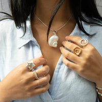 Person wearing a necklace with a shell pendant and multiple rings on a light blue shirt - Seol Gold