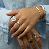 Close-up of hands wearing multiple rings with spiral, star, and other designs on a light blue fabric background - Seol Gold