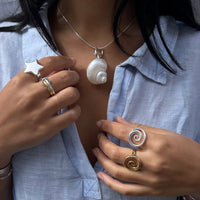 Person wearing a necklace with a shell pendant and multiple rings on a light blue shirt - seolgold