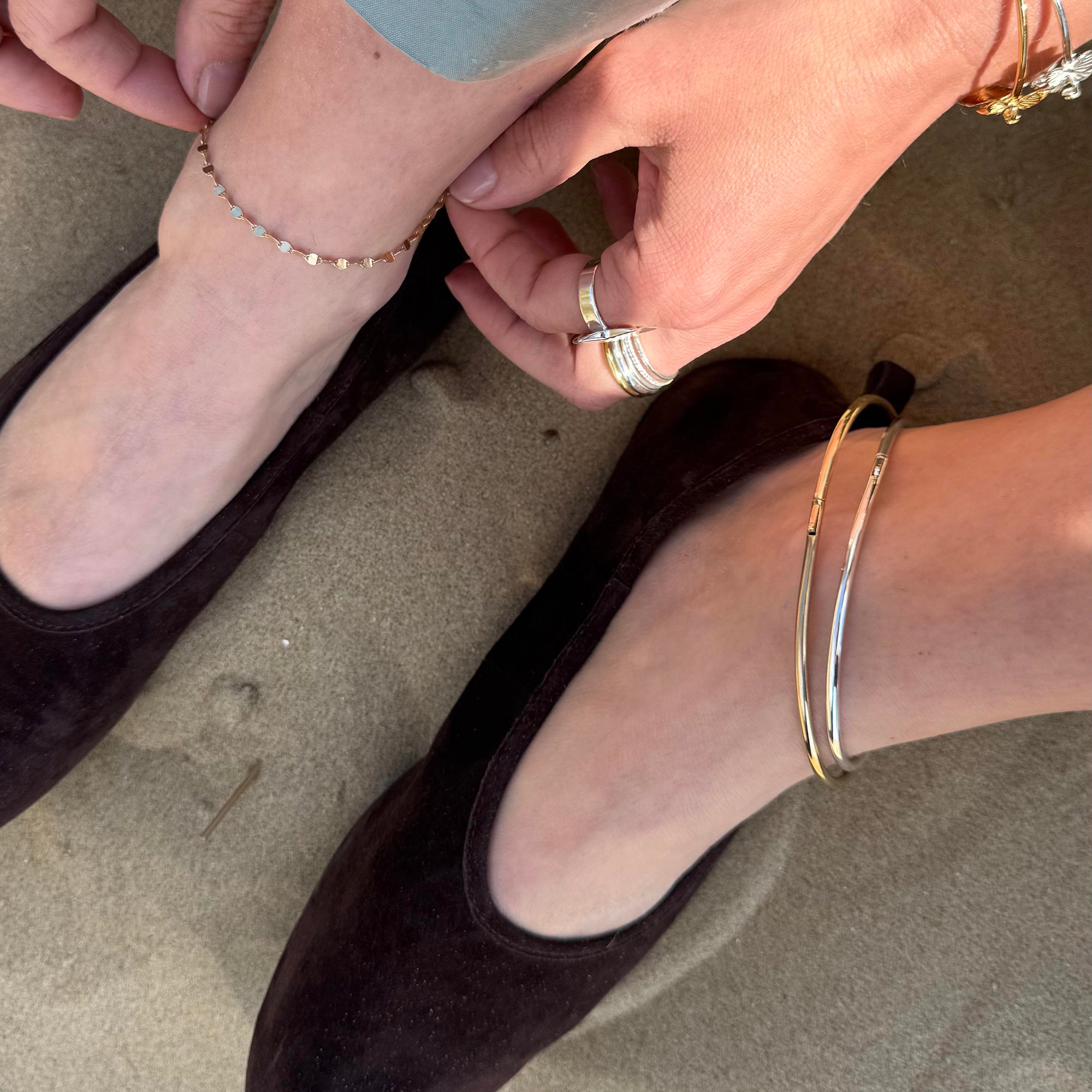 Close-up of feet wearing black shoes with gold and silver ankle jewelry on a neutral background - seolgold