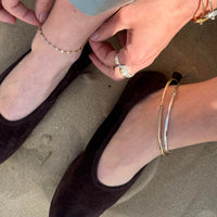 Close-up of feet wearing black shoes with gold and silver ankle jewelry on a neutral background - seolgold