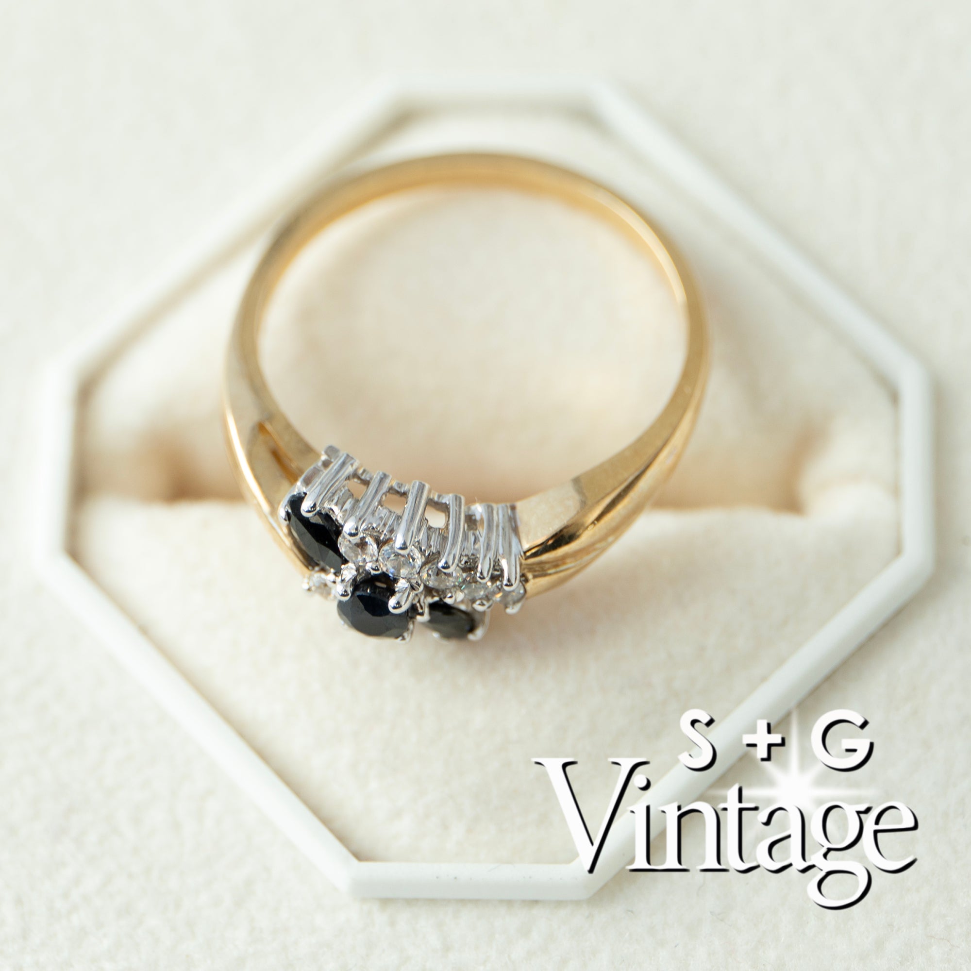 Gold ring with black and white gemstones on a hexagonal white stand, branded 'S + G Vintage' - seolgold